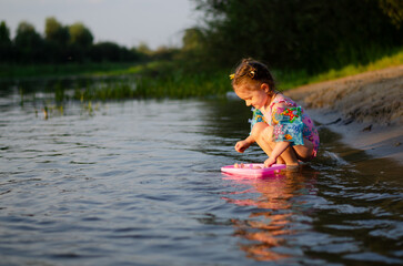 Fototapeta premium Little 3-year-old girl in inflatable arm floaties and swimsuit holding a doll by the river