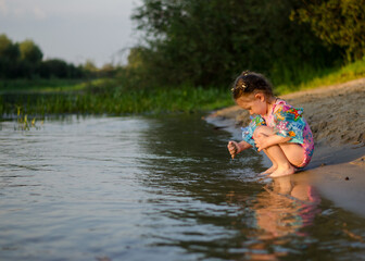 Fototapeta premium Little 3-year-old girl in inflatable arm floaties and swimsuit holding a doll by the river
