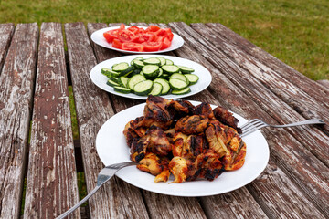 A picnic table with three white plates containing grilled meat, tomatoes and cucumbers.