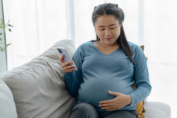 Young asian pregnant woman sitting on sofa using her mobile phone