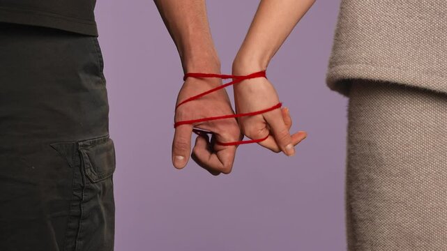 Man and woman hands tied together, tangled with red thread, love and codependency concept, isolated on purple studio background, close up shot.