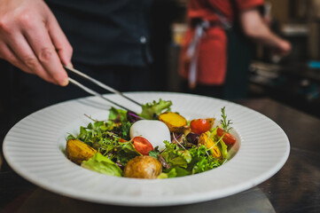 A chef uses tongs to arrange a fresh salad on a plate. The vibrant mix includes greens, cherry tomatoes, and roasted potatoes, topped with a poached egg, showcasing culinary artistry.
