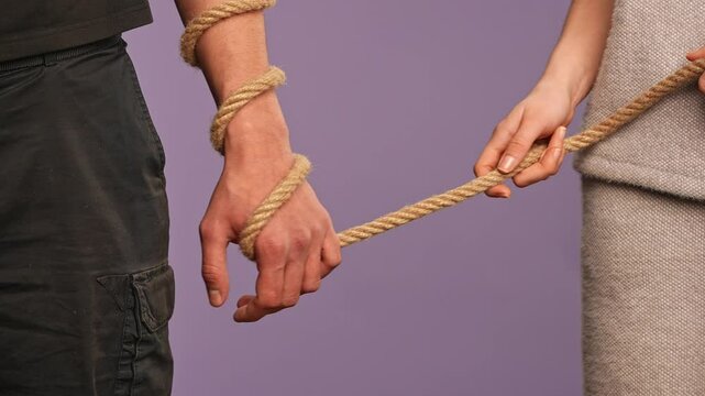 Man hand tied with rope, woman holding the rope as leash, abusive dominated feministic relationships concept, isolated on purple studio background, close up shot.