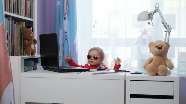 A cheerful cute baby girl, wearing sunglasses, sits at her workdesk next to a laptop and notebook. Surrounded by toys and books