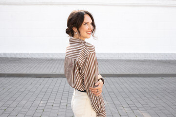 Fototapeta premium Portrait of young beautiful brunette woman wearing silk striped brown shirt. Girl standing outdoor on a city street against a white wall. Trendy casual summer or spring outfit. Street fashion.
