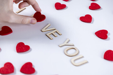 person arranging wooden letters to spell love you with a red felt heart on a white background surrounded by scattered red hearts for Valentine's Day