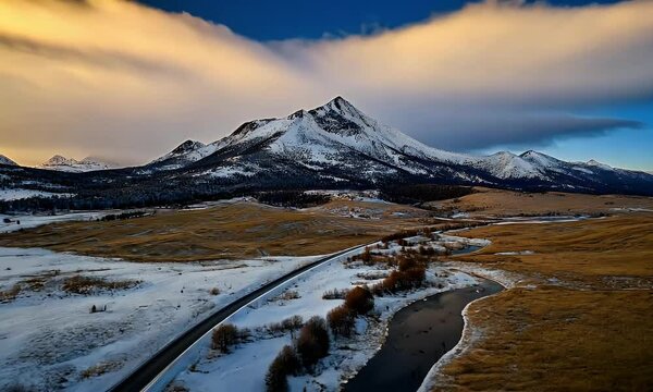 Snow-Capped Mountain Peak Surrounded by Open Fields and a Serene Winding Road at Sunset with Vibrant Cloud Formations in a Tranquil Winter Landscape