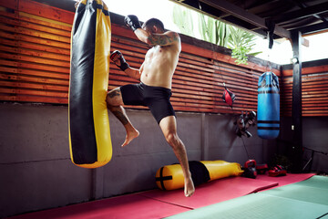 Shirtless martial arts male fighter practicing flying knee kick on punching bag in home gym
