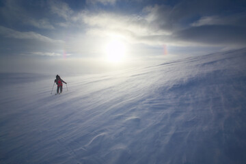 A lone skier crosses a huge snowy pass under bright sunshine and cloudy skies.