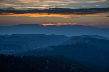 Beautiful Sunset With Blue Colored Trees In The Foreground And Foggy Hills In The Background. Orange And Yellow Sky With Clouds