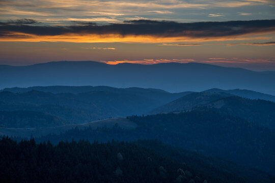 Beautiful Sunset With Blue Colored Trees In The Foreground And Foggy Hills In The Background. Orange And Yellow Sky With Clouds