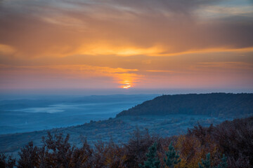 Beautiful Sunset With Blue Colored Trees In The Foreground And Foggy Hills In The Background. Orange And Yellow Sky With Clouds