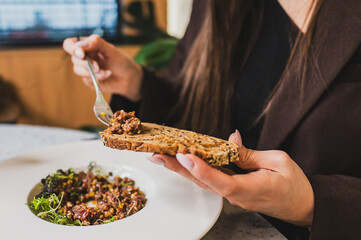 A close-up of a hand holding a slice of grain bread topped with a savory spread, while the other hand holds a fork near a bowl of mixed salad. A cozy dining setting.