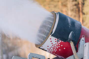 Close-up of a red snow cannon spraying artificial snow with frosty details on the machine, against a blurred forest background.