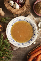 Chicken bone broth in white plate with blue ornaments on a wooden table with fresh vegetables