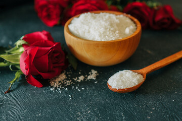 Wooden bowl filled with white bath salt, surrounded by vibrant red roses and scattered petals on a dark textured surface, creating a romantic and natural spa-like ambiance