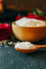 Wooden bowl filled with white bath salt, surrounded by vibrant red roses and scattered petals on a dark textured surface, creating a romantic and natural spa-like ambiance
