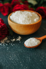 Wooden bowl filled with white bath salt, surrounded by vibrant red roses and scattered petals on a dark textured surface, creating a romantic and natural spa-like ambiance