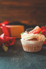 Close-up of a cupcake topped with whipped cream, strawberry, and mint, placed on a dark textured background with red roses and a gift box in the background, creating a festive mood