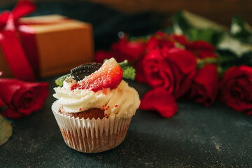 Close-up of a cupcake topped with whipped cream, strawberry, and mint, placed on a dark textured background with red roses and a gift box in the background, creating a festive mood