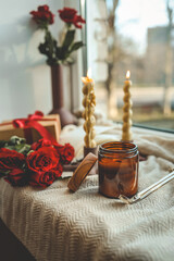Two twisted candles burning in modern holders by a windowsill, accompanied by a red ribbon gift box, red roses, and a vase with roses, creating a cozy ambiance with natural light