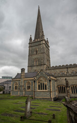 Obraz premium Saint Columb's Cathedral with a Gothic spire and weathered gravestones under an overcast sky, Londonderry, Northern Ireland