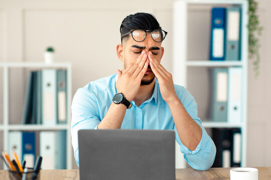 Millennial Arab employee rubbing tired irritated eyes, working with laptop too much, exhausted from online job at office. Young Eastern businessman feeling fatigue from looking at screen