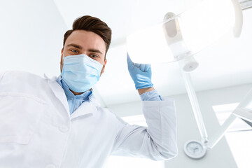 Fototapeta premium Male dentist in medical face mask turning on lamp, greeting patient in dental chair, looking at camera, copy space. Young man stomatologist beginning treatment, turning on light, closeup