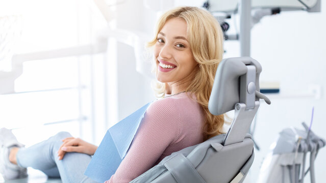 Smiling and relaxed young woman sitting at dental chair in modern clinic, waiting for her dentist. Happy attractive blonde lady attending luxury dental clinic, bacl view, copy space