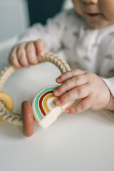 Baby holding a silicone rainbow teether with braided handle and pastel-colored details. Perfect for sensory play and teething relief. Modern and functional design