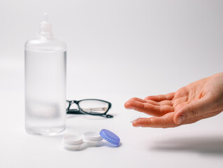 Hand holding a transparent contact lens next to lens case, solution bottle, and glasses on a white background. Concept of eye care, vision correction, and optical hygiene
