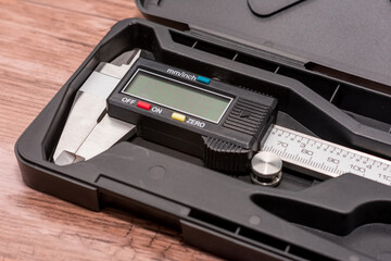 Digital caliper lies in a black case on a wooden surface, featuring a black and silver design with a clear digital screen. Combination of precise engineering and natural wood creates visual contrast