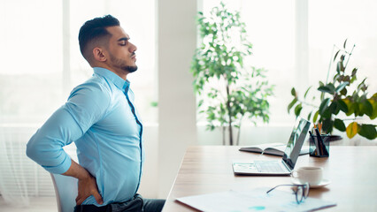 Young Arab businessman having acute back pain, massaging aching muscles, sitting at desk in modern...