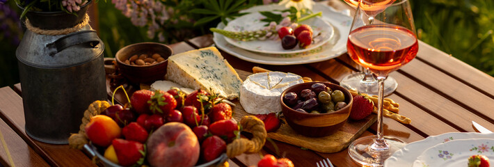 Banner. Two glasses of rose wine, flowers in a vase, silverware, fruits. Sunset, golden hour, atmospheric candlelight. Romantic table decor for a loving couple on blooming meadow with purple lupines
