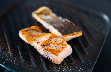 There are two pieces of broiled salmon steaks in square black frying pan. Steam is billowing over freshly cooked fish fillet.
