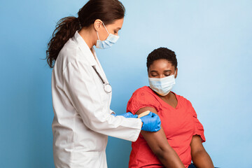 Fototapeta premium Antiviral Immunization, Covid-19 Vaccination. Black Female Getting Vaccinated, Doctor Sticking Adhesive Bandage After Vaccine Injection On Patient's Arm Over Blue Background. Studio Shot