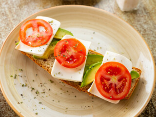 Mediterranean open-faced sandwich with fresh tomato, avocado slices, and soft cheese on crispy toast, topped with herbs, perfect for healthy snack