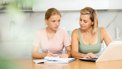 Obraz premium Teenage girl student engrossed in studies, sitting at table with laptop and diligently writing notes in notebook guided by positive friendly female tutor