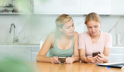 Middle-aged woman and teenage daughter are sitting in kitchen with phones in their hands. Girl...
