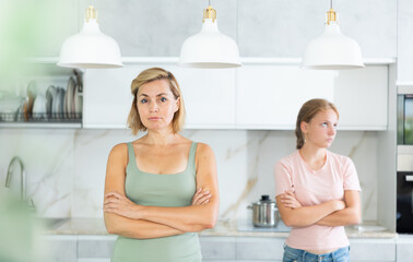 Portrait of upset woman standing in home kitchen on background of offended frustrated teenage daughter standing behind. Family conflicts and communication problems concept