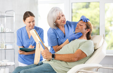 Interested adult man looking at mirror while skilled female cosmetologist showing results of facial procedure in medical esthetic office. Female assistant filling out patient medical record