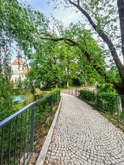 A scenic view of a stone-paved path curved between railings, surrounded by lush green vegetation, with a glimpse of an elegant building in the background under a clear sky.
