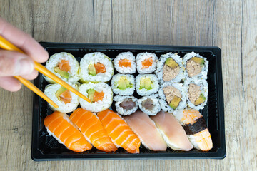 From above anonymous hand with chopsticks eating assorted sushi rolls served on tray on wooden restaurant table with copy space.