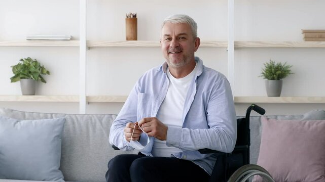 A mature Caucasian man in a wheelchair adjusts his mask while seated in a cozy, bright living room.
