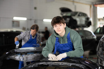 Young professional auto body technician in blue overalls and casual green hoodie, engaged in restoring damaged cars, expertly sanding removed bumper with sandpaper to smooth surface before painting