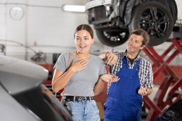 Happy young female customer receiving remote key fob for repaired car from smiling mature auto mechanic in automotive repair shop after successful service..