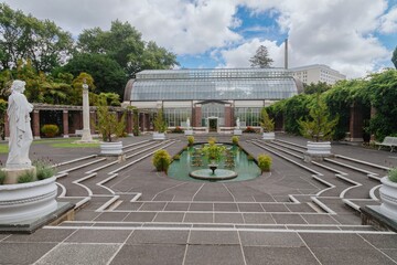 Peaceful garden courtyard with a glasshouse, fountain, and statues. Visitors stroll through the...