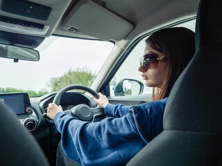 A woman is driving a car and looking at the road. She is wearing a blue sweater. Young inexperienced driver behind driving wheel. Cost of insurance for new driver. Travel and tourism