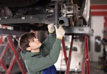 Confident young auto mechanic in blue overalls standing under car lifted on hydraulic lift in...