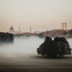 Fototapeta premium A Misty Morning View Of Munich Old Town From The English Garden With Fog Draping The Landscape And Hazy Silhouettes Of Historic Architecture